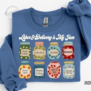 May include: Indigo blue sweatshirt with the text "Labor & Delivery is My Jam" in white. The sweatshirt features a graphic of nine jars with different stages of labor and delivery, such as "Early Labor", "Contractions", "Dilation", and "Placenta Delivery".