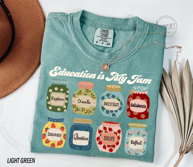 May include: Light green t-shirt with the text "Education is My Jam" printed on the front. The text is surrounded by colorful illustrations of jars with different words on them, such as "Explore", "Create", "Investigate", "Collaborate", "Innovate", "Question", "Solve", and "Reflect".