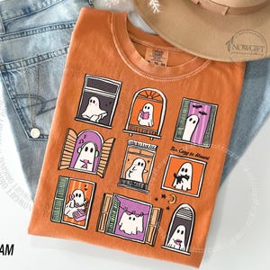May include: Orange t-shirt featuring a design of ghosts in windows. The ghosts are engaged in various activities, such as reading and playing music. The text "Too Cozy to Haunt" is printed on the shirt. The shirt is folded on top of blue jeans.