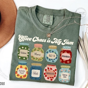 May include: A moss green t-shirt featuring the text "Office Chaos is My Jam" and jam jar illustrations. The jars are labelled with words such as "Visitor" and "Attendance". A silver necklace is draped across the shirt.