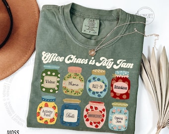 Op de afbeelding: Mosgroen t-shirt met de tekst "Office Chaos is My Jam" en illustraties van jampotten. De potten hebben labels zoals "Visitor" en "Attendance". Een zilveren ketting ligt over het shirt.