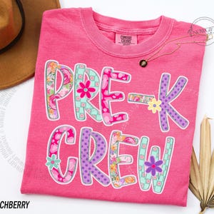 May include: A bright pink t-shirt with the words "PRE-K CREW" in a colorful, floral design. The letters are outlined in white and feature various patterns, including flowers and polka dots. The shirt has a crew neck and short sleeves.