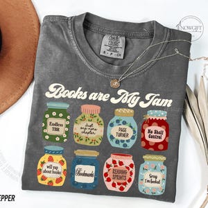 May include: A grey t-shirt featuring the text "Books are My Jam" and illustrations of jam jars. Each jar has a different label, such as "Endless TBR" and "Page Turner." A silver necklace with a small pendant is on the shirt.