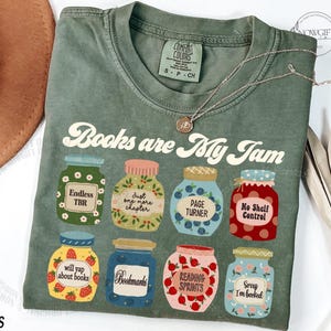 May include: Moss green t-shirt featuring the text "Books are My Jam" and illustrated jam jars with labels like "Endless TBR" and "Page Turner". A silver necklace and a brown hat are also visible in the image.