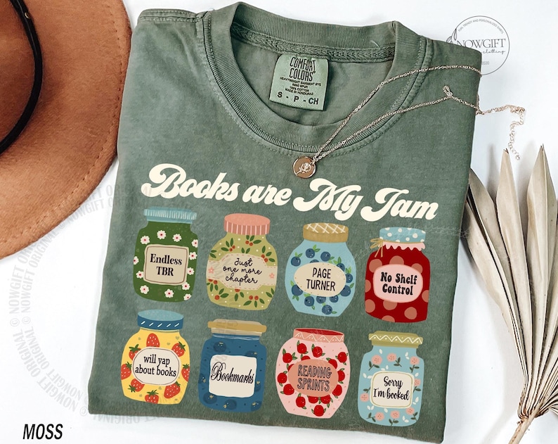 May include: Moss green t-shirt featuring the text "Books are My Jam" and illustrated jam jars with labels like "Endless TBR" and "Page Turner". A silver necklace and a brown hat are also visible in the image.