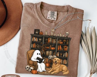 Op de afbeelding: Een espresso-kleurig t-shirt met een afbeelding van een geest die een boek leest in een fauteuil naast een golden retriever. De scène bevat een boekenplank vol boeken, pompoenen, kaarsen en schedels. Het shirt heeft een Comfort Colors label.