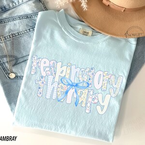 Pu&ograve; includere: Una maglietta azzurra con la scritta "respiratory therapy" in un design patchwork. Le lettere sono bianche con motivi floreali e a quadri, e un fiocco blu. La maglietta &egrave; piegata sopra un paio di jeans azzurri e un cappello beige.