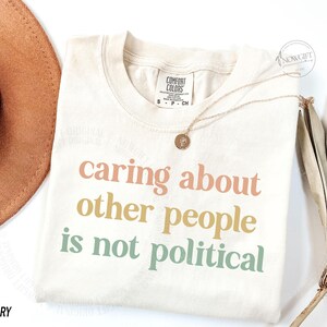 May include: A cream colored t-shirt with the text "caring about other people is not political" printed in pastel colors. The text is in a bold, sans-serif font.