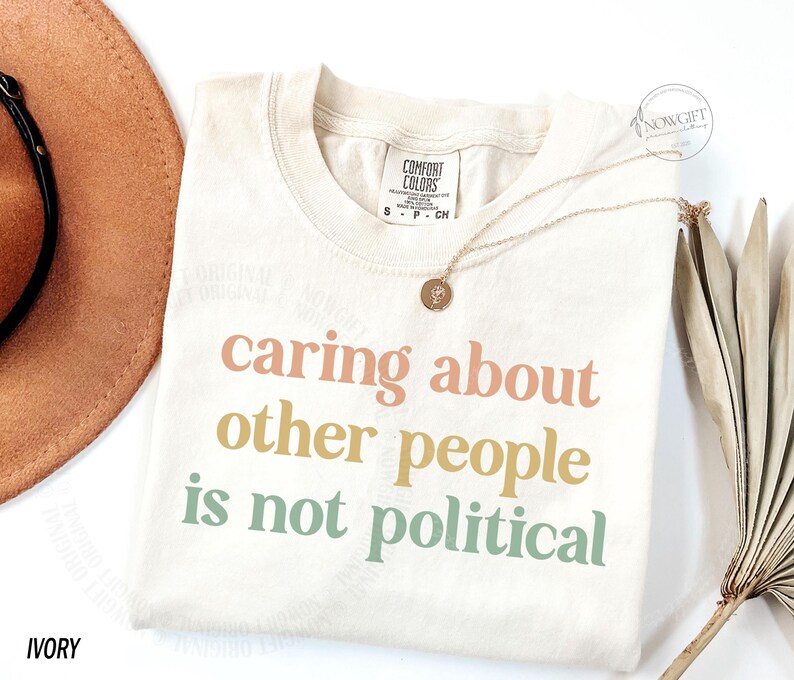 May include: A cream colored t-shirt with the text "caring about other people is not political" printed in pastel colors. The text is in a bold, sans-serif font.