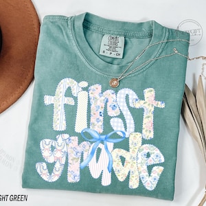 May include: A light green t-shirt with the words "First Grade" appliqued in various floral and patterned fabrics. A blue bow is below the words. A gold necklace with a pendant is on the shirt.