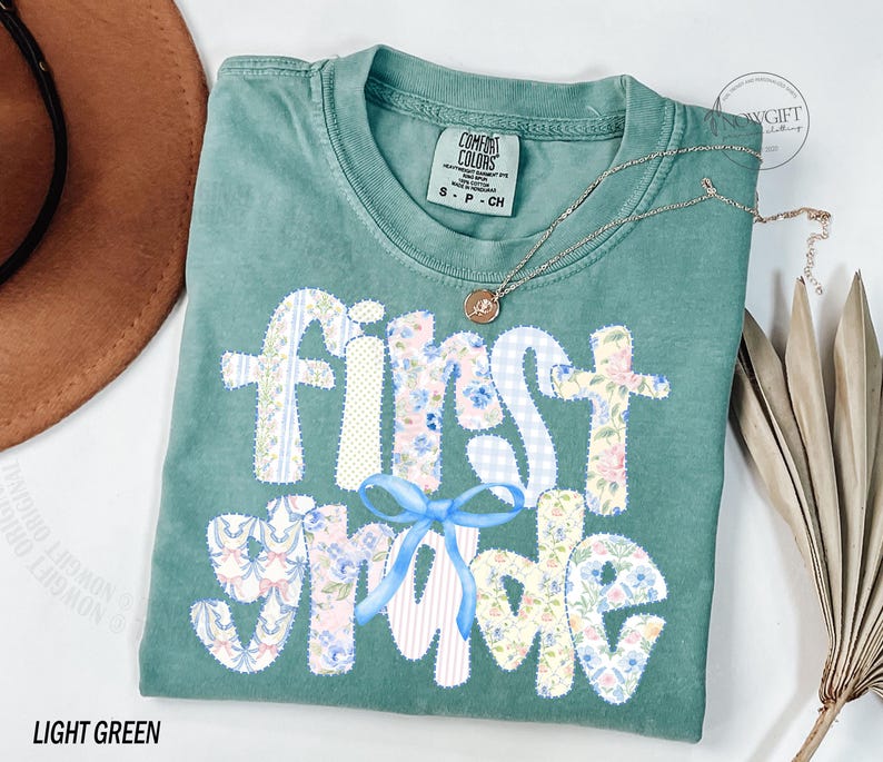 May include: A light green t-shirt with the words "First Grade" appliqued in various floral and patterned fabrics. A blue bow is below the words. A gold necklace with a pendant is on the shirt.