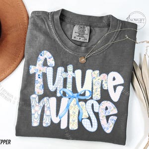 May include: A gray t-shirt with the words "future nurse" in a patchwork design with floral and gingham patterns. A light blue bow accents the word "nurse". A gold necklace with a pendant is on the shirt.