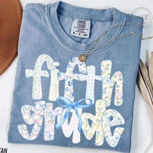May include: Blue jean Comfort Colors t-shirt with "fifth grade" in a patchwork design. The letters are made of floral and gingham patterns, with a blue bow accent. A gold necklace with a pendant is draped on the shirt.