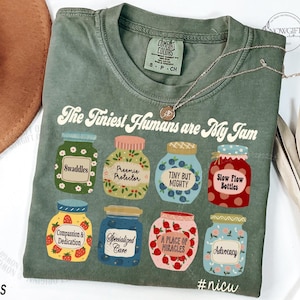 Puede incluir: Camiseta Comfort Colors verde musgo con el texto "The Tiniest Humans are My Jam". La camiseta presenta ilustraciones de tarros coloridos con etiquetas como "Swaddles" y "Slow Flow Bottles". Un delicado collar descansa sobre la camiseta. El hashtag #nicu es visible.