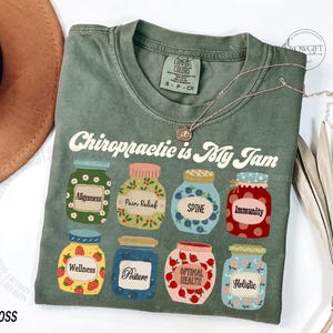 May include: A moss green t-shirt with the phrase "Chiropractic is My Jam" printed in white. The shirt features illustrations of jars labeled with chiropractic terms like "Alignment" and "Immunity". A silver necklace rests on the shirt.