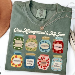 May include: Moss green t-shirt with the phrase "Case Management is My Jam" and colorful jar graphics. Each jar represents a quality like "Empathy" and "Advocacy". A silver necklace with a pendant is draped on the shirt.