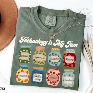 May include: A moss green t-shirt featuring the text "Technology is My Jam" and jar illustrations with words like "Innovate" and "Collaborate." A gold necklace is placed on the shirt.