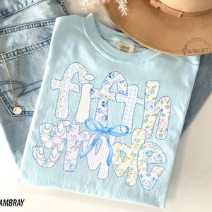 May include: Light blue t-shirt with the words "Fifth Grade" in a patchwork design. The letters are decorated with floral and gingham patterns, and a blue bow is centered below the words. The word "Chambray" is printed at the bottom.
