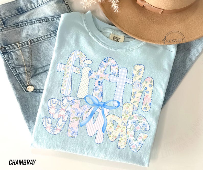 May include: Light blue t-shirt with the words "Fifth Grade" in a patchwork design. The letters are decorated with floral and gingham patterns, and a blue bow is centered below the words. The word "Chambray" is printed at the bottom.