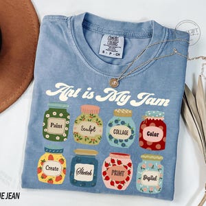 Op de afbeelding: Een blauw jeans T-shirt met de tekst "Art is My Jam" erop gedrukt. Het shirt heeft een ontwerp van zeven kleurrijke potten met verschillende kunstgerelateerde woorden erop, waaronder "Verf", "Beeldhouwen", "Collage", "Kleur", "Cre&euml;ren", "Schetsen", "Drukken" en "Digitaal".