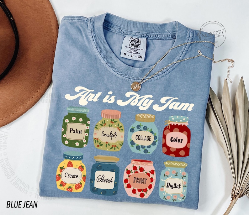 Op de afbeelding: Een blauw jeans T-shirt met de tekst "Art is My Jam" erop gedrukt. Het shirt heeft een ontwerp van zeven kleurrijke potten met verschillende kunstgerelateerde woorden erop, waaronder "Verf", "Beeldhouwen", "Collage", "Kleur", "Cre&euml;ren", "Schetsen", "Drukken" en "Digitaal".