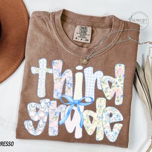 Puede incluir: Camiseta color espresso con las palabras "third grade" en un dise&ntilde;o de patchwork. Las letras presentan patrones florales y de cuadros, con un lazo azul claro. Un collar plateado con un colgante est&aacute; drapeado sobre la camiseta.