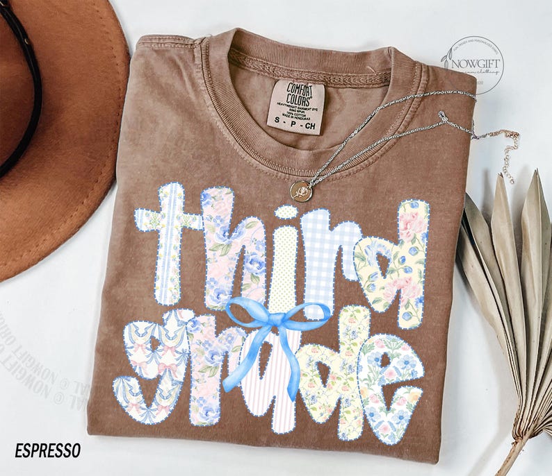 Puede incluir: Camiseta color espresso con las palabras "third grade" en un dise&ntilde;o de patchwork. Las letras presentan patrones florales y de cuadros, con un lazo azul claro. Un collar plateado con un colgante est&aacute; drapeado sobre la camiseta.