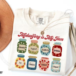 May include: Ivory-colored t-shirt with the phrase "Midwifery is My Jam" in red script. Below, illustrated jars feature words like "Education," "Support," and "Respect." A gold necklace and a hat are also visible.