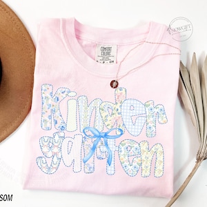 Pu&ograve; includere: Una maglietta Comfort Colors rosa chiaro con la scritta "Kindergarten" in un design patchwork con motivi floreali e a quadretti. Un fiocco blu &egrave; sotto la parola. Una collana d'oro e un cappello marrone sono accanto alla maglietta. La parola "Blossom" &egrave; in basso.