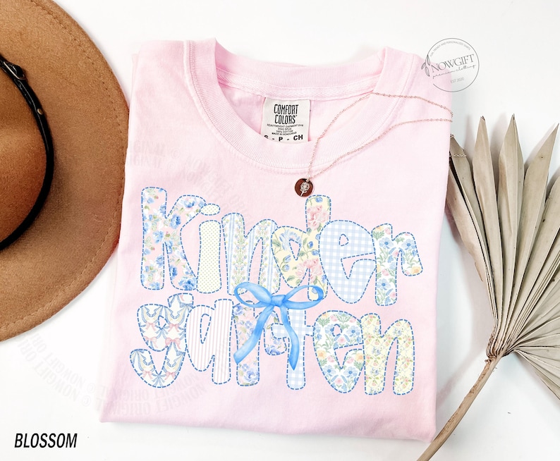 Pu&ograve; includere: Una maglietta Comfort Colors rosa chiaro con la scritta "Kindergarten" in un design patchwork con motivi floreali e a quadretti. Un fiocco blu &egrave; sotto la parola. Una collana d'oro e un cappello marrone sono accanto alla maglietta. La parola "Blossom" &egrave; in basso.