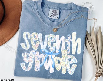 Op de afbeelding: Een blauw Comfort Colors T-shirt met de woorden "Seventh Grade" in een patchwork-ontwerp. De letters zijn versierd met bloemen- en streepjespatronen. Een blauwe strik is onder de woorden gecentreerd. Een gouden ketting met een hanger ligt op het shirt.