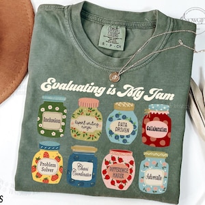 May include: A green t-shirt with the text "Evaluating is My Jam" printed on it. The text is in a retro font and is surrounded by illustrations of jars with different labels, such as "Inclusion", "Data Driven", and "Problem Solver".