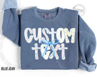 Op de afbeelding: Een blauw spijkerbroekkleurig sweatshirt met de woorden "CUSTOM TEXT" in een patchwork-ontwerp. De letters zijn wit met bloemen- en ruitpatronen. In het midden is een blauw lint geknoopt. De mouwen zijn opgerold.