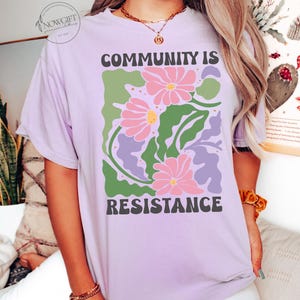 May include: A lavender t-shirt with a graphic design of pink flowers and green leaves. The text "COMMUNITY IS RESISTANCE" is printed in black letters on the shirt.