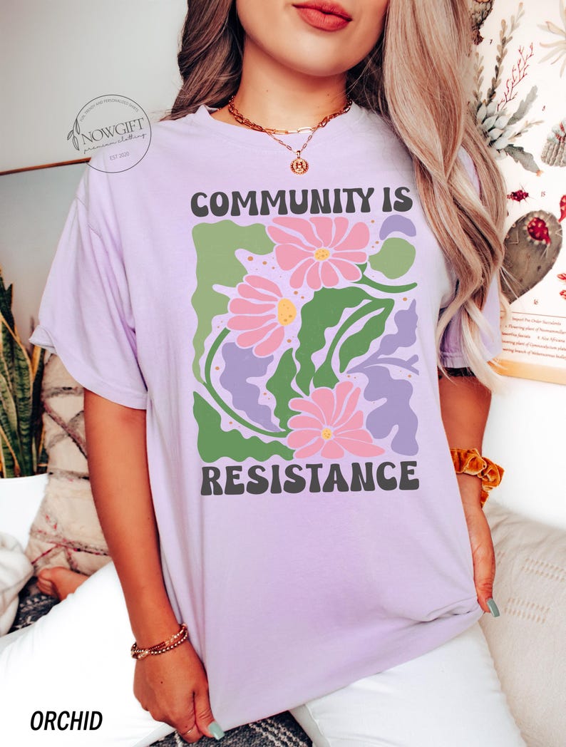 May include: A lavender t-shirt with a graphic design of pink flowers and green leaves. The text "COMMUNITY IS RESISTANCE" is printed in black letters on the shirt.