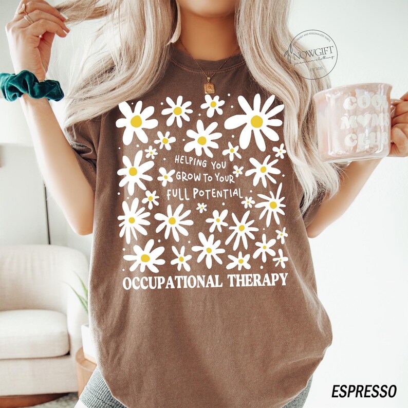 May include: Espresso-colored t-shirt with a daisy floral design and the text "Helping You Grow To Your Full Potential Occupational Therapy." The shirt has a crew neck and short sleeves. The shirt is a relaxed fit.