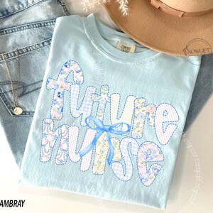 May include: A light blue t-shirt with the words "Future Nurse" appliqued in a patchwork style. The letters are in various floral and patterned fabrics, with a blue bow. The shirt is folded on top of a pair of blue jeans.