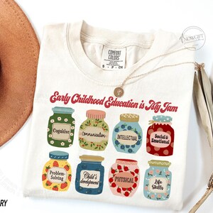 Puede incluir: Una camiseta de color crema con el texto "Early Childhood Education is My Jam" en rojo. La camiseta presenta un gr&aacute;fico de nueve frascos con diferentes etiquetas, que incluyen "Cognitive", "Communication", "Intellectual", "Social & Emotional", "Problem-Solving", "Child's Development", "Physical" y "Life Skills".