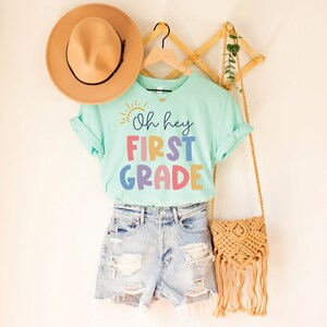 May include: A light blue t-shirt with the text "Oh hey FIRST GRADE" in rainbow colors. The shirt is hanging on a wooden hanger with a straw hat and a woven bag.