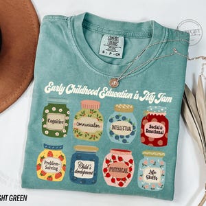 Puede incluir: Camiseta verde claro con el texto "Early Childhood Education is My Jam" impreso en la parte delantera. El texto est&aacute; rodeado de ilustraciones de frascos con diferentes etiquetas, que incluyen "Cognitive", "Communication", "Intellectual", "Social & Emotional", "Problem-Solving", "Child's Development", "Physical" y "Life Skills".