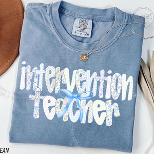 May include: A blue jean Comfort Colors t-shirt with the words "Intervention Teacher" in a floral and gingham pattern. A light blue bow is centered below the word "Intervention". A gold necklace with a pendant is draped across the shirt.