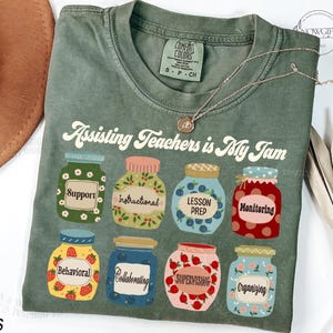 May include: A green t-shirt with the text "Assisting Teachers is My Jam" printed on it. The text is in a retro font. There are eight jars with different designs and text printed on them. The jars are filled with different coloured items, such as strawberries, blueberries, and flowers.