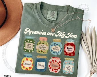 Op de afbeelding: Mosgroen t-shirt met de tekst "Preemies are My Jam" en illustraties van potten met diverse labels. De labels bevatten "Slow Flow Bottle", "Tiny Miracles" en "Caffeinated".