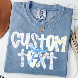 Könnte beinhalten: Ein blaues Comfort Colors T-Shirt mit der Aufschrift "CUSTOM TEXT" in einem Blumen- und Vichy-Muster, akzentuiert mit einem blauen Band. Eine goldene Halskette mit Anhänger liegt auf dem Shirt.