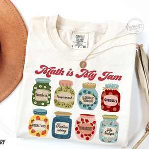 May include: A white t-shirt with the text "Math is My Jam" in red and a graphic of jars with different math concepts, such as numbers, measurement, algebraic thinking, geometry, operations, problem solving, probability, and data analysis.