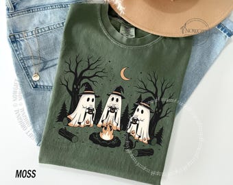 Op de afbeelding: Een mosgroen t-shirt met een grafische print van drie geesten met zwarte heksenhoeden, zittend rond een kampvuur in een bos bij nacht. De geesten houden mokken vast en een maansikkel is zichtbaar in de lucht boven de donkere bomen.