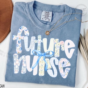 May include: A blue jean Comfort Colors t-shirt with the words "Future Nurse" in white, floral, and gingham patches. A light blue bow is centered below the word "future". A gold necklace with a pendant is draped across the shirt.