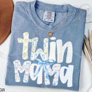 Puede incluir: Camiseta azul vaquero Comfort Colors con las palabras "twin mama" en un dise&ntilde;o de patchwork. Las letras est&aacute;n decoradas con patrones florales y de rayas, y un lazo azul. Un collar dorado est&aacute; drapeado sobre la camisa.