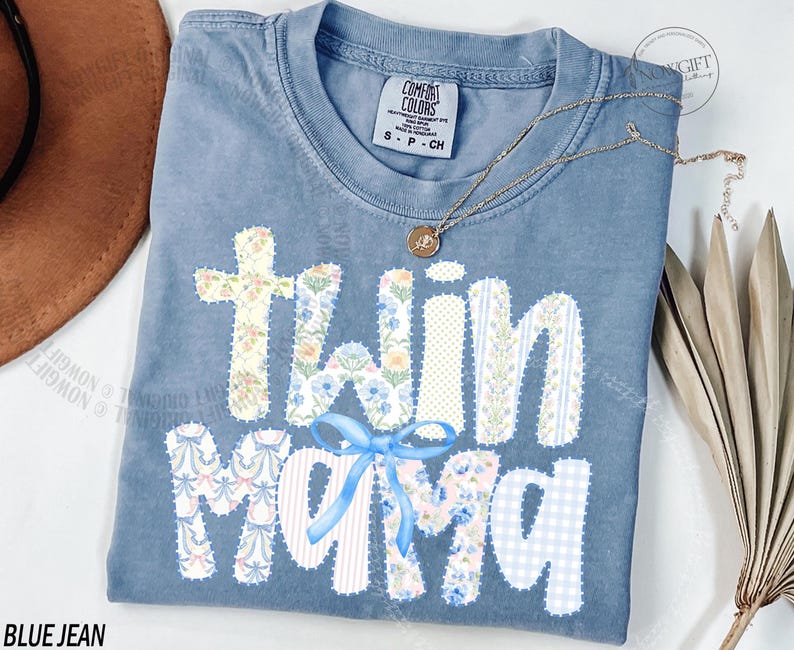 Puede incluir: Camiseta azul vaquero Comfort Colors con las palabras "twin mama" en un dise&ntilde;o de patchwork. Las letras est&aacute;n decoradas con patrones florales y de rayas, y un lazo azul. Un collar dorado est&aacute; drapeado sobre la camisa.