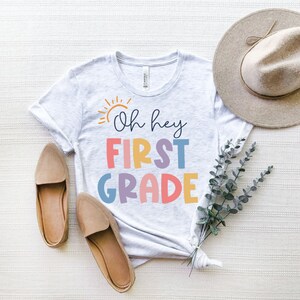 May include: A white t-shirt with the text "Oh hey first grade" in rainbow colors. The shirt is laid out on a white textured surface with a brown hat and brown shoes.